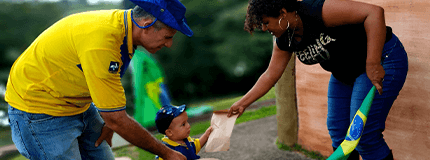 Carteiro uniformizado, acompanhado de uma criança com uniforme dos Correios, entregando uma carta a uma mulher.