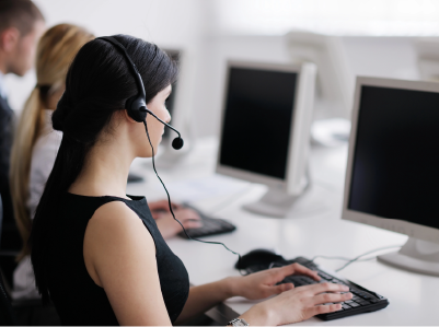 Visão lateral de uma moça atendente branca de cabelos lisos pretos presos e camiseta preta com fone e microfone no ouvido e as mãos no teclado sentada em frente ao computador