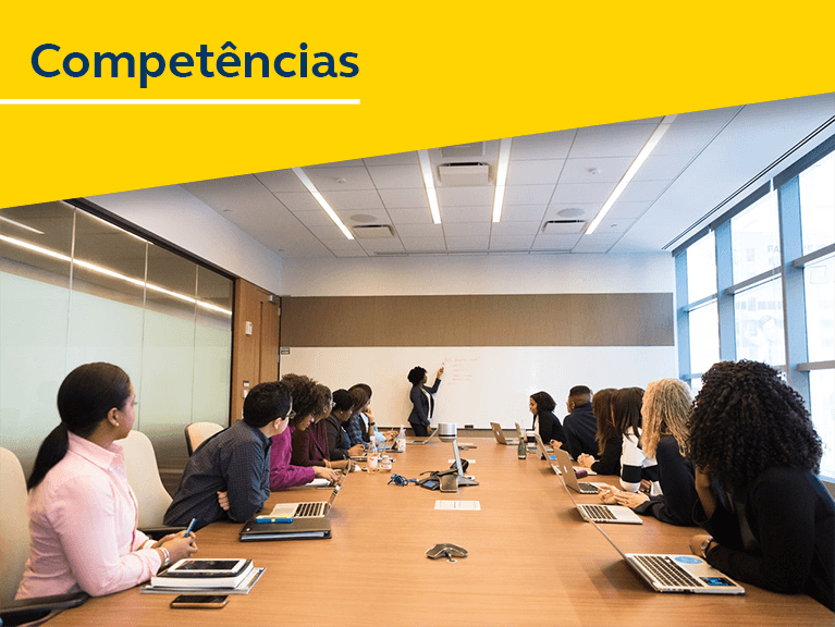 Pessoas reunidas ao redor de uma mesa, com notebooks a sua frente, com os olhares voltados a uma pessoa a frente de uma quadro branco realizando apontamentos.