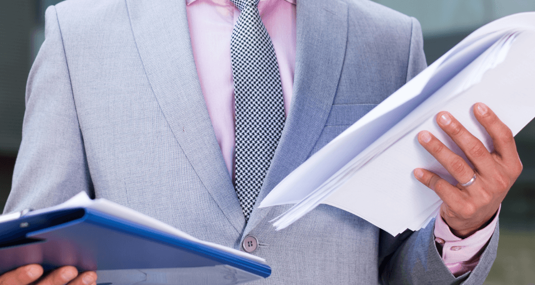 Busto de homem usando terno azul claro, camisa rosa claro, e gravata cinza claro, segurando pasta de documentos com capa azul escuro na mão direita e na mão esquerda, algumas folhas de papel branco