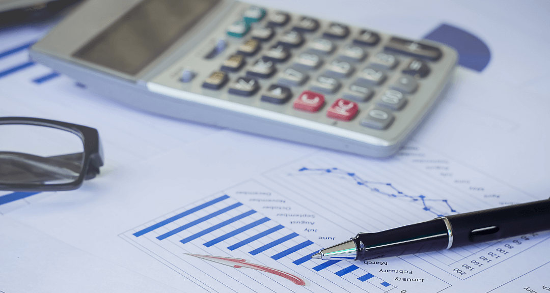 Calculadora cinza sobre uma mesa com algumas folhas de papel com desenhos de alguns gráficos, caneta preta e parte de um óculos preto