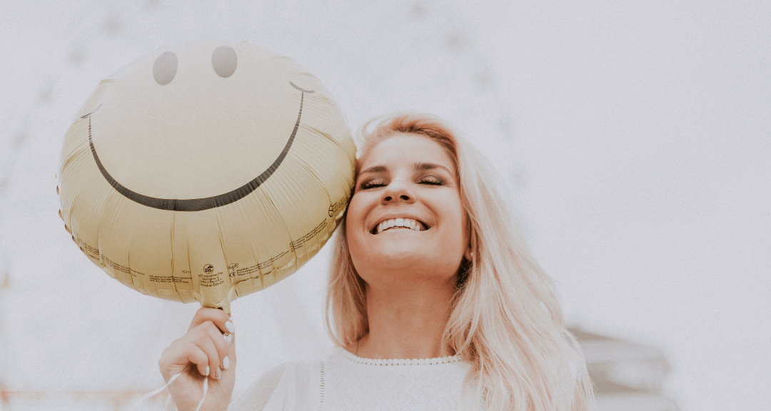 Mulher loira, com camisa branca, segurando balão com um sorriso