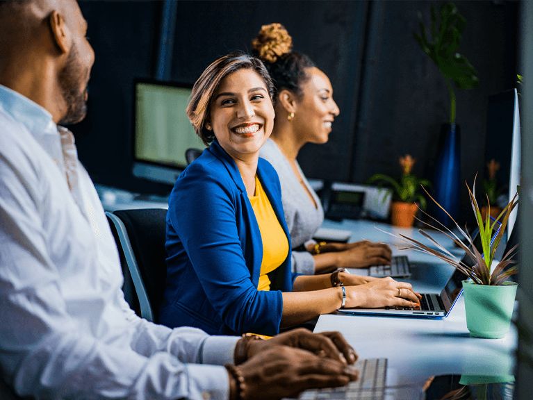 Pessoas trabalhando utilizando notebook, com expressões de interação, uma sorrindo para o outro.