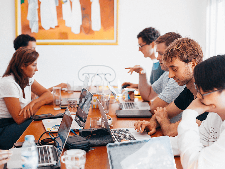 Várias pessoas sentadas à mesa, uma de frente para a outra com notebooks.