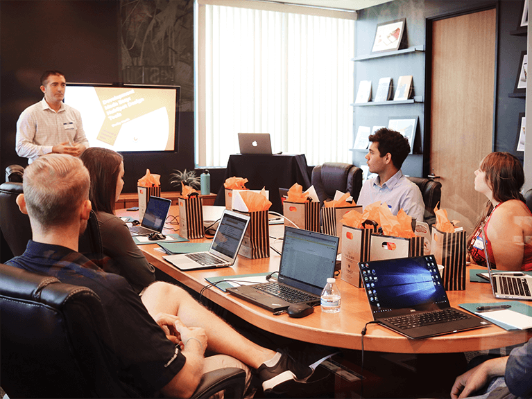 pessoas reunidas ao redor de uma mesa, cada um com notebook, com os olhares voltados para a pessoa que encontra-se em pé.