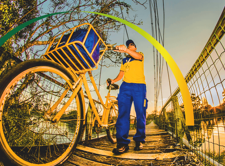 Carteiro, de uniforme azul e amarelo, atravessa uma ponte de madeira empurrando uma bicicleta