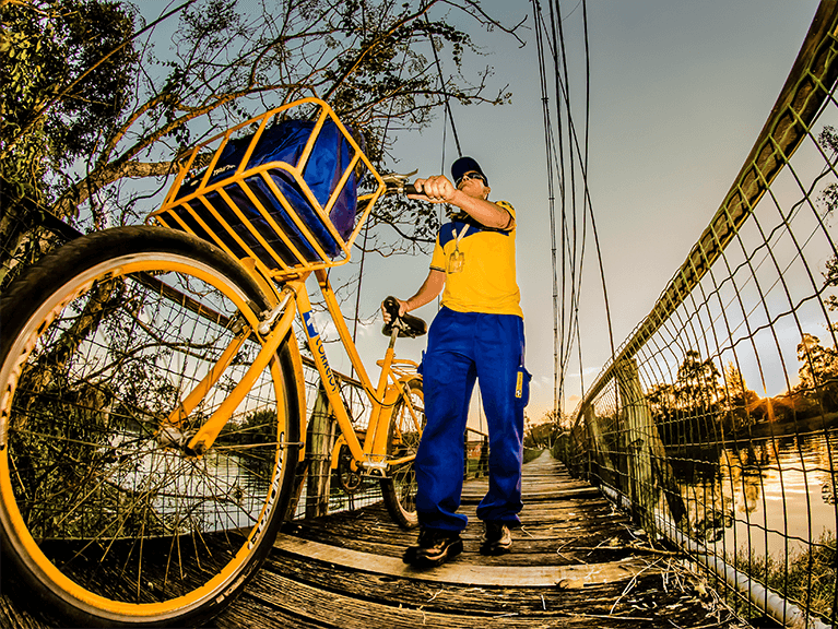 Universalização Carteiro com bicicleta dos Correios passando por uma ponte localizada acima de um rio