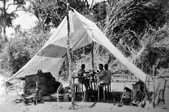 Comissões Foto em preto e branco de pessoas embaixo de uma tenda improvisada. Ao fundo uma mata.