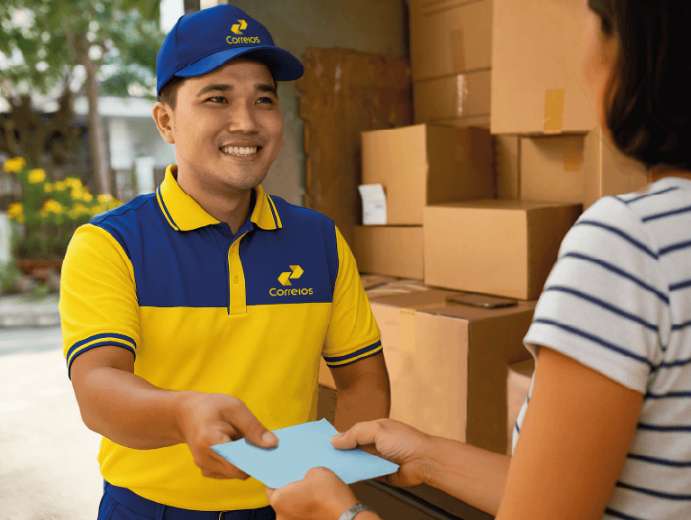 Imagem de carteiro uniformizado entregando um papel para uma moça de costas e camiseta listrada e caixas de encomenda em um caminhão estacionado na rua.