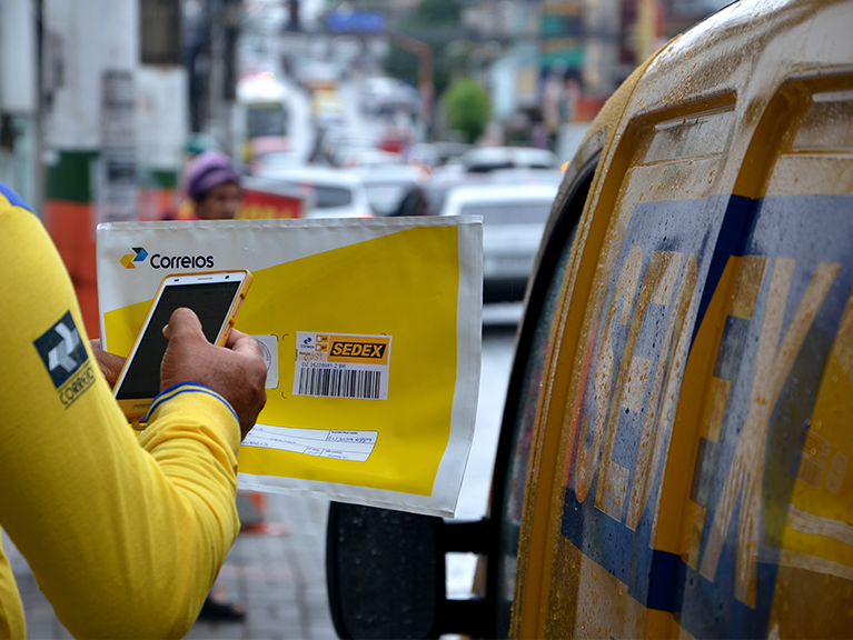 Sedex Hoje Carteiro fotografando o código de barras de uma encomenda em frente a um carro amarelo dos correios com a descrição do SEDEX. Frase do card: Sedex Hoje