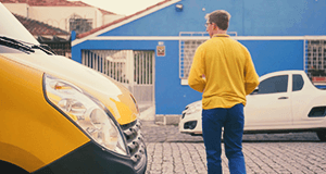 Foto de um carteiro atravessando a rua em frente ao carro amarelo dos Correios. Ao fundo um carro branco estacionado em frente a uma casa azul Frase do card: Sedex 10