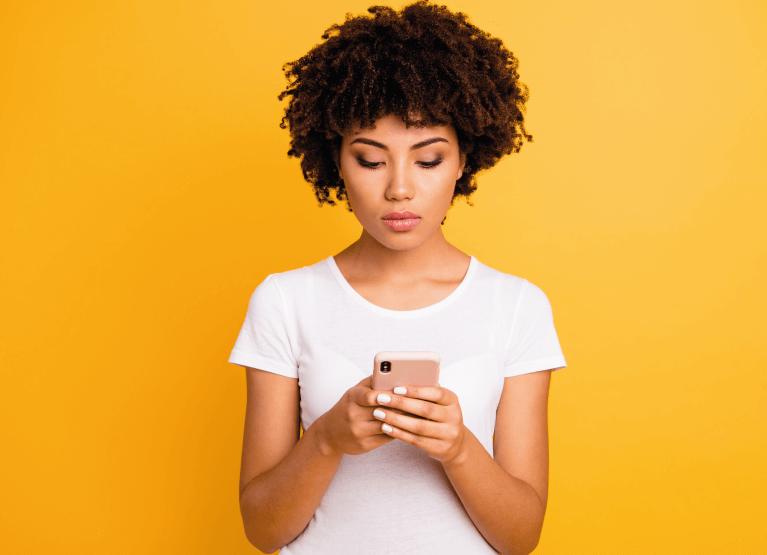 Fundo amarelo, mulher em pé, blusa branca, segura e olha fixamente o celular com as duas mãos