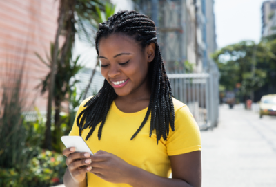 Mulher negra, cabelos com tranças, blusa amarela, sorrindo, segura o celular com as duas mãos. Ao fundo uma imagem embaçada de uma rua