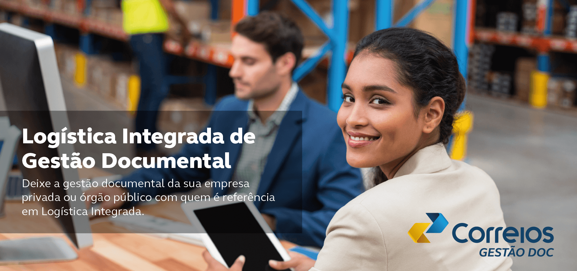 Foto de mulher morena e terno bege olhando pra trás com tablet na mão em uma mesa com um homem branco de terno azul manuseando um computador e prateleiras de um armazém atrás. Frase: Logística Integrada de Gestão Documental. Deixe a gestão documental da sua empresa privada ou órgão público com quem é referência em Logística Integrada.