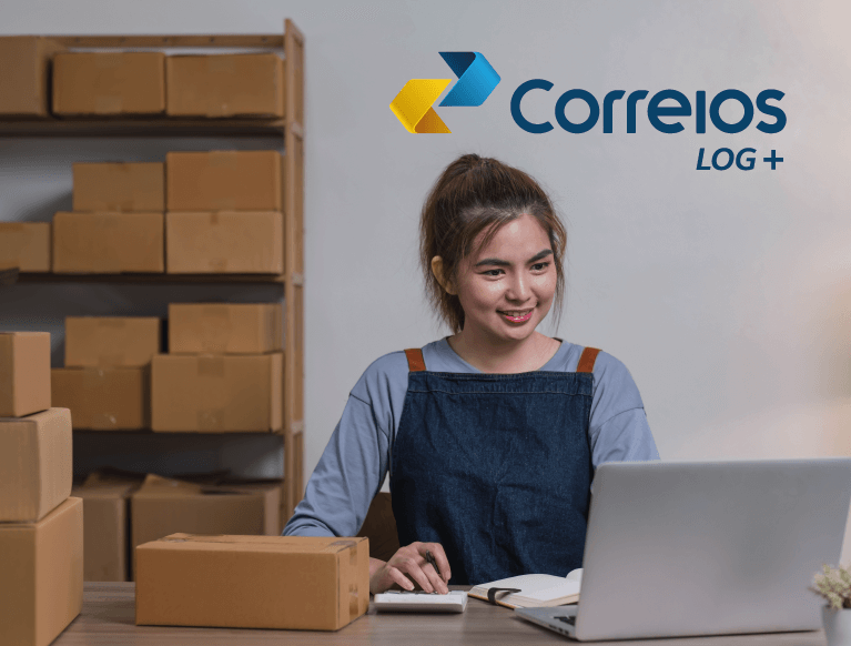 Mulher morena, sentada em frente a uma mesa e um notebook, sorrindo. Ao fundo um armário com caixas de papelão